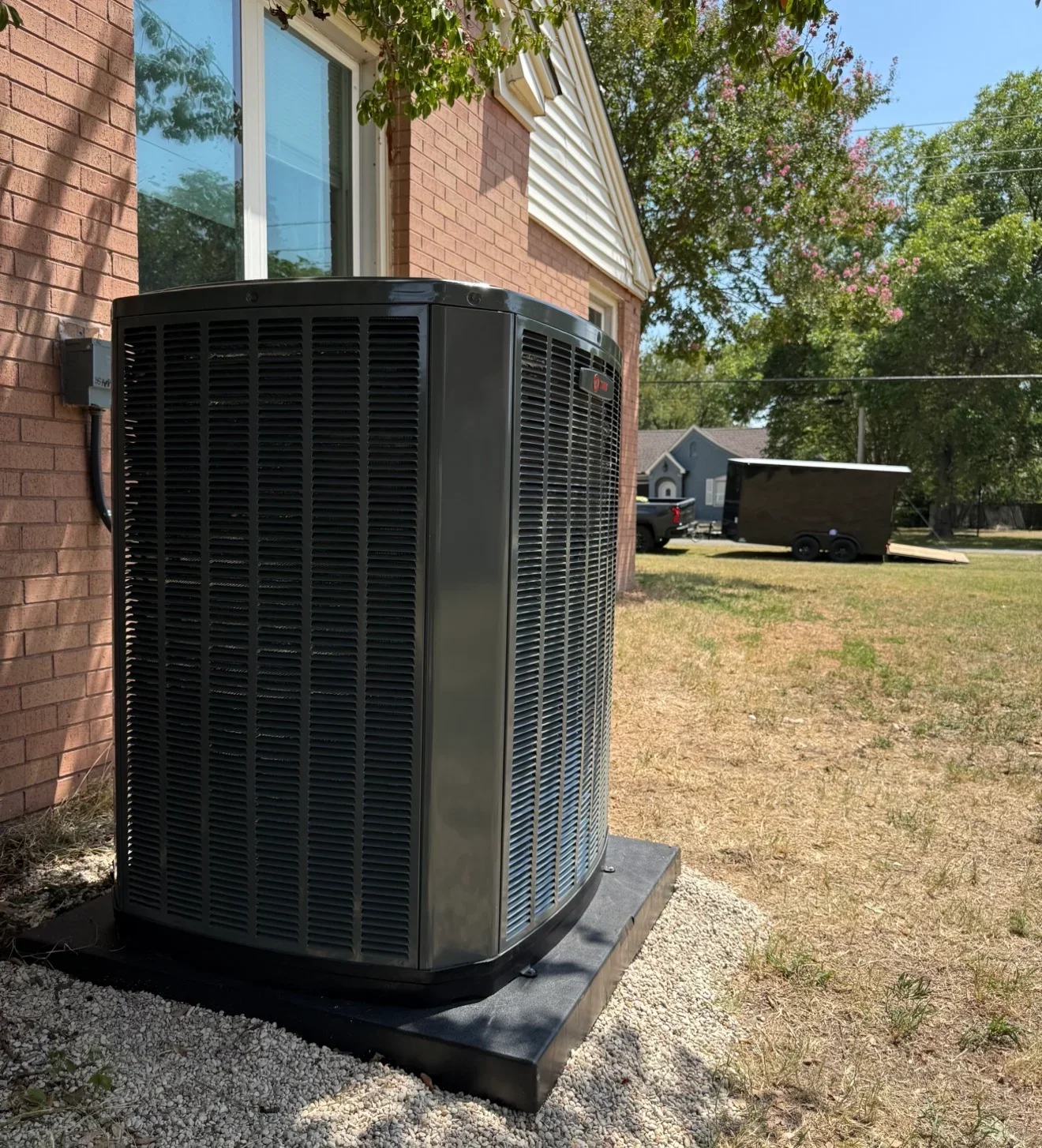 Dark gray outdoor AC condenser on black composite pad and white gravel beside red brick wall