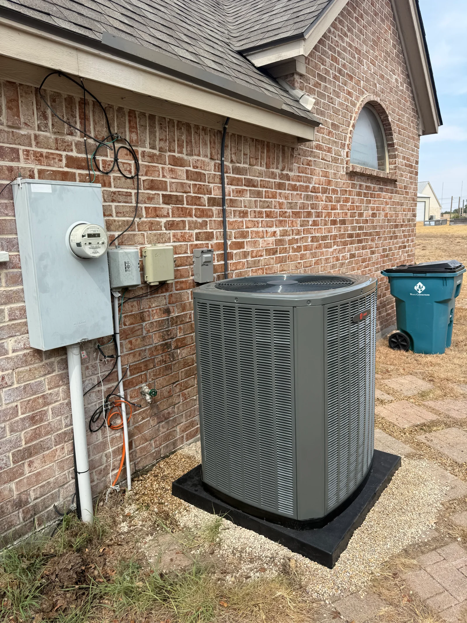 Gray outdoor AC condenser on black pad with white gravel beside red brick wall, electrical disconnect and conduit
