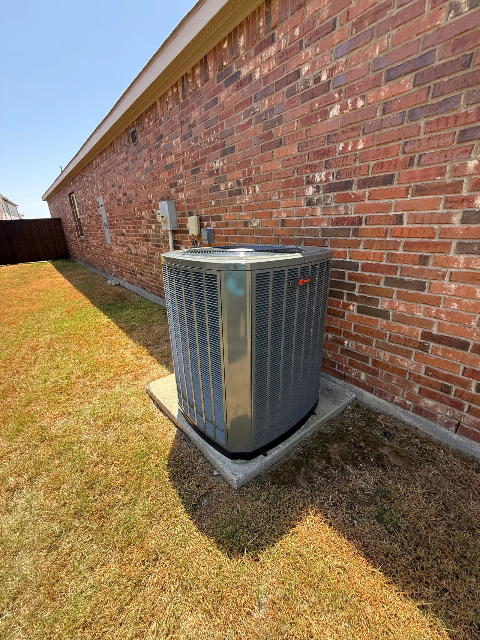 Trane outdoor AC condenser on concrete pad against red brick wall with louvered metal panels
