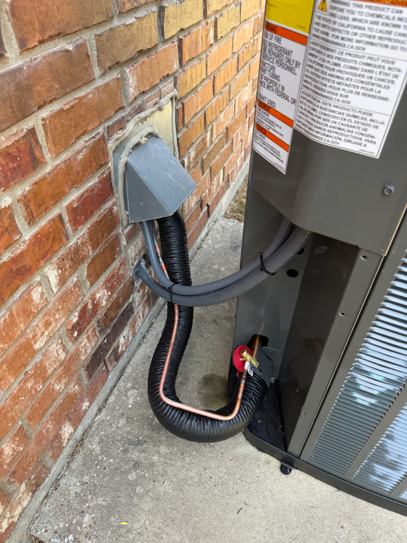 Outdoor AC condenser with black insulated line set, copper refrigerant lines, and gray wall sleeve against brick wall